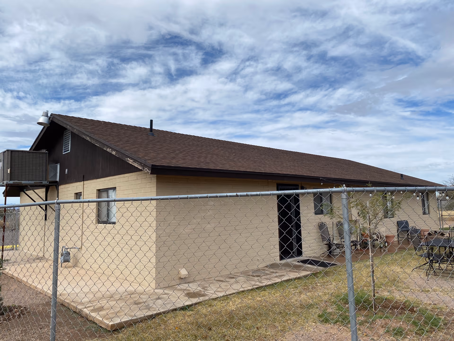 Single-story beige brick assisted living building with a brown roof seen behind a chain-link fence under a cloudy sky.