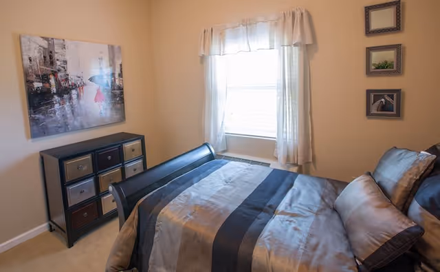 A bedroom with a bed covered in a striped comforter in shades of gray and blue, a window with sheer white curtains, a small dresser with multiple drawers, and framed artwork on the walls.