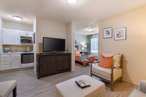 Open interior of an assisted living apartment showing a seating area with an armchair and ottoman, a TV on a dresser, a kitchenette to the left, and a bed in the adjoining bedroom.