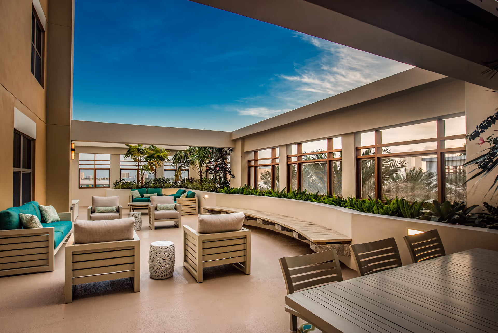 A spacious, covered outdoor patio area with modern beige and teal cushioned seating, including sofas and armchairs, surrounded by plants and palm trees. The area features a long curved bench along the wall and a dining table with chairs in the foreground. The sky is clear with some clouds visible through the open roof structure.