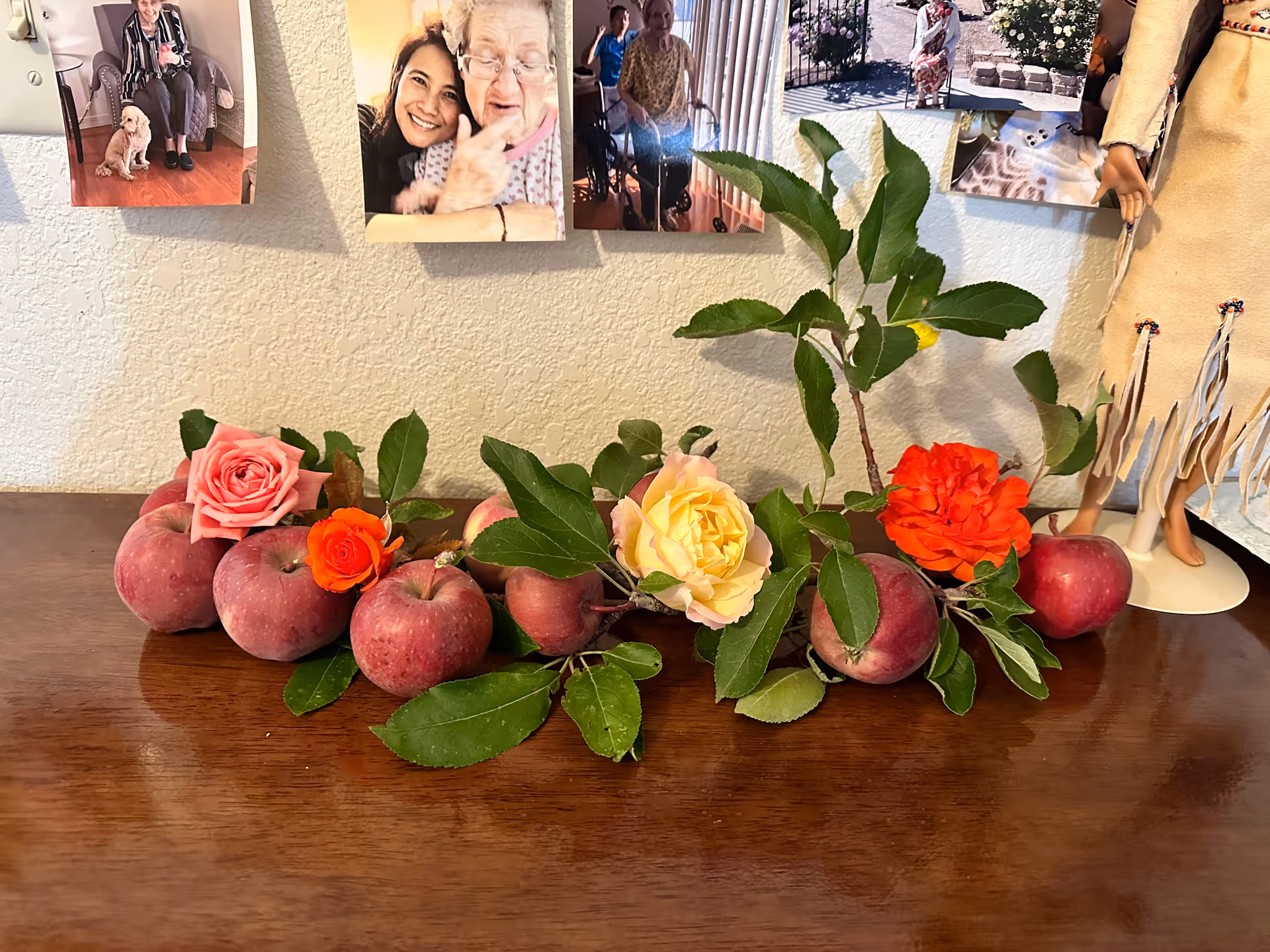 A decorative arrangement on a wooden surface featuring red apples, green leaves, and colorful roses in pink, orange, and yellow. Behind the arrangement, there are several photographs pinned to a textured wall, showing elderly people and a dog. To the right, part of a doll dressed in traditional clothing is visible.