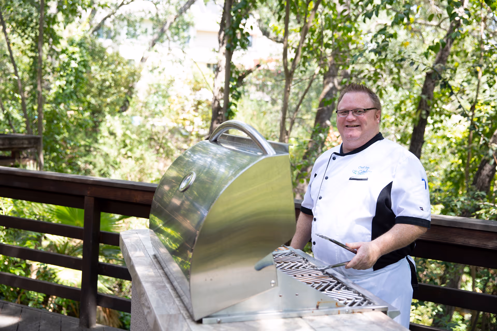 A man wearing a white chef's coat with black accents is standing outdoors on a wooden deck next to a stainless steel grill. He is holding grilling tongs and smiling. The background features green trees and foliage.