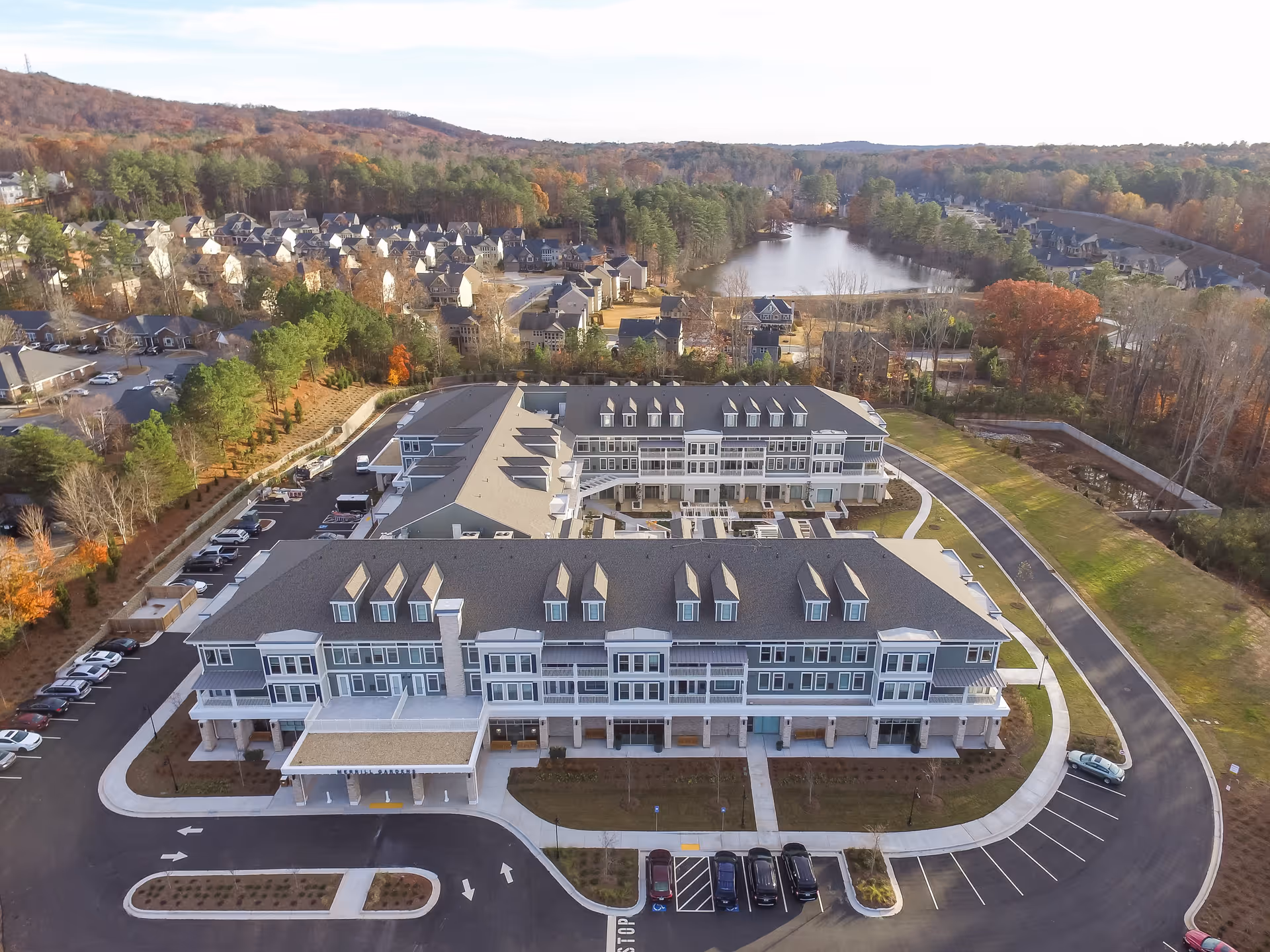 Aerial view of Merrill Gardens at Woodstock, showing a large multi-story senior living facility surrounded by parking lots, roads, and landscaped areas. In the background, there are residential houses, trees with autumn foliage, and a small lake.