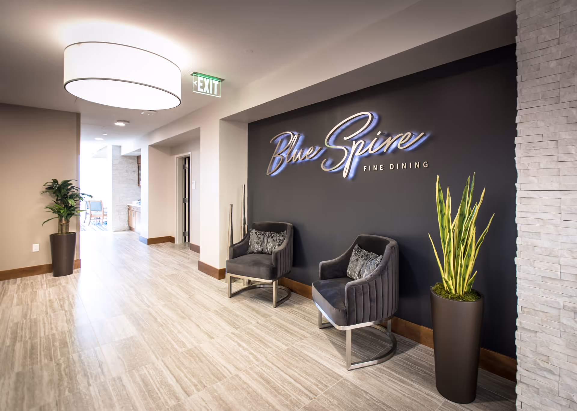 A hallway in a senior living facility with a sign on a dark wall that reads 'Blue Spire Fine Dining.' Two gray upholstered chairs with patterned cushions are placed against the wall, flanked by tall potted plants. The floor is tiled with light-colored stone, and a large round ceiling light fixture illuminates the space. An exit sign is visible above a doorway leading to another area.