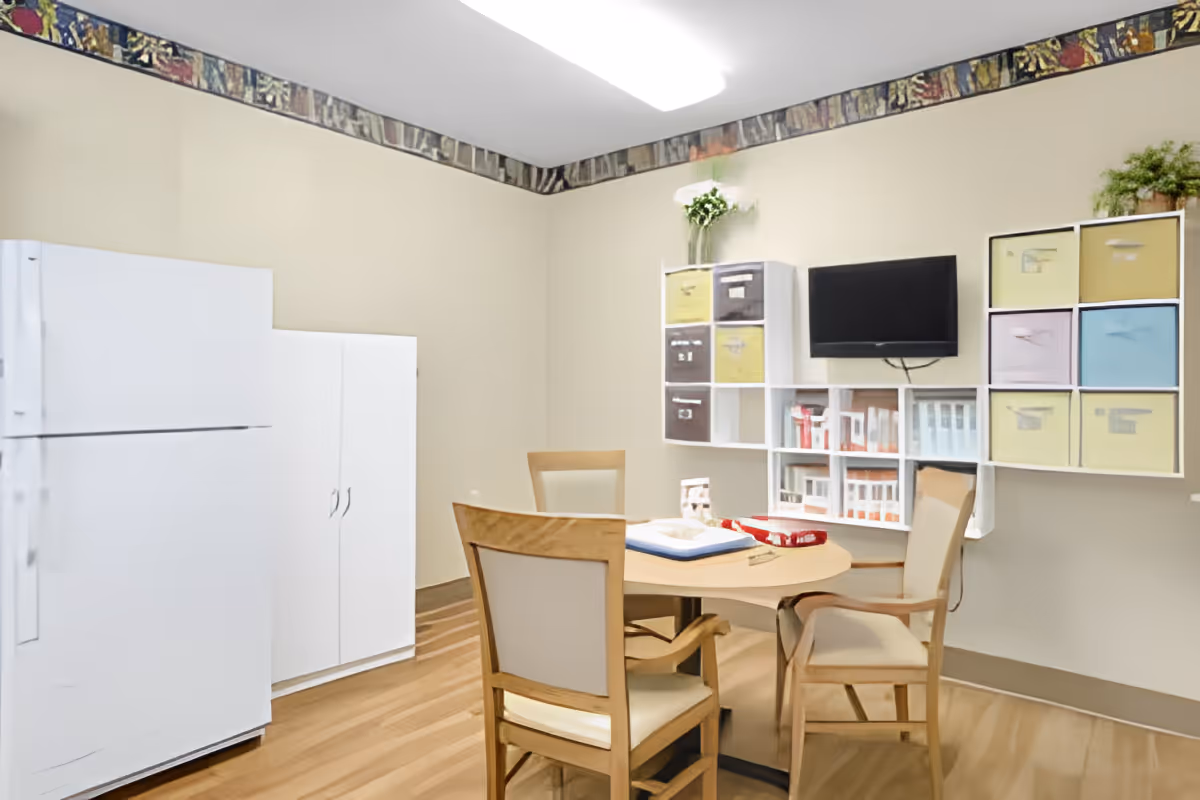 A small room with a round wooden table surrounded by four wooden chairs with beige cushions. On the table are some board games and a book. The room has light beige walls with a decorative border near the ceiling. There is a white refrigerator and a white cabinet against one wall. On another wall, there are white cubby shelves with colorful storage bins and a small flat-screen TV mounted above them. A small plant is placed on top of one of the shelves.