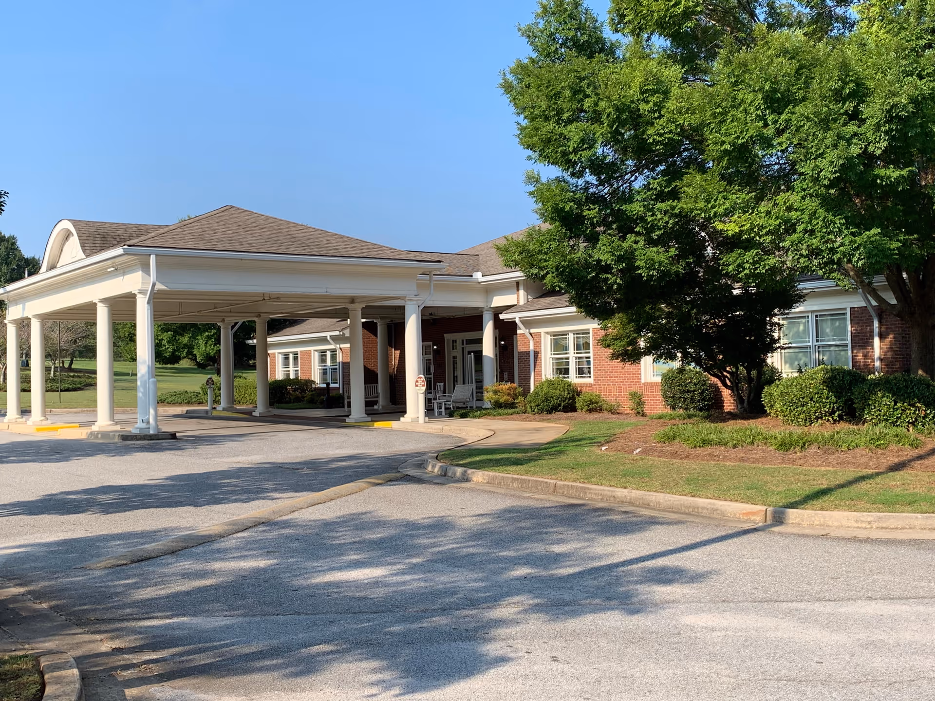 Exterior view of Brightmoor Nursing Center showing a covered entrance with white columns, a brick building, green bushes, and a large tree under a clear blue sky.