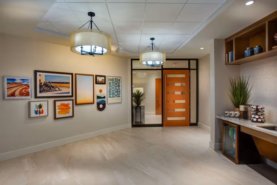 A modern interior space with beige walls and tiled floor, featuring a wall decorated with various framed artworks. Two large round pendant lights hang from the ceiling. On the right side, there is a countertop with a coffee machine, mugs, a small refrigerator, and a potted plant. A wooden door with horizontal glass panels leads to another room.