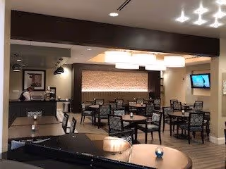 Interior view of a dining area in a senior living facility with multiple tables and chairs arranged neatly. The room has modern lighting fixtures, a flat-screen TV mounted on the wall, and a counter area where a staff member is standing. The decor includes neutral tones and a textured wall panel behind the counter.