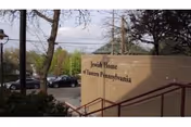 Outdoor view of a brick wall with the sign 'Jewish Home of Eastern Pennsylvania' near a parking lot with several cars and trees in the background.