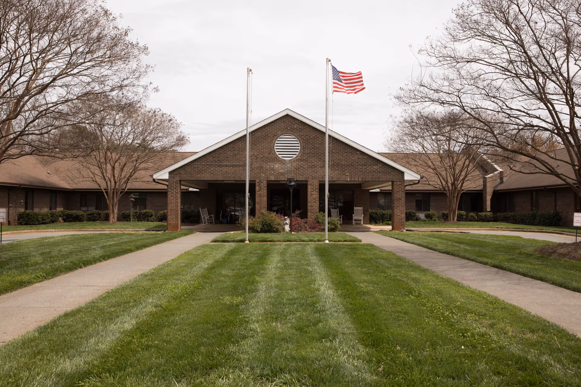 Front entrance of a single-story brick senior living building with an American flag on a flagpole, walkways and a manicured lawn.