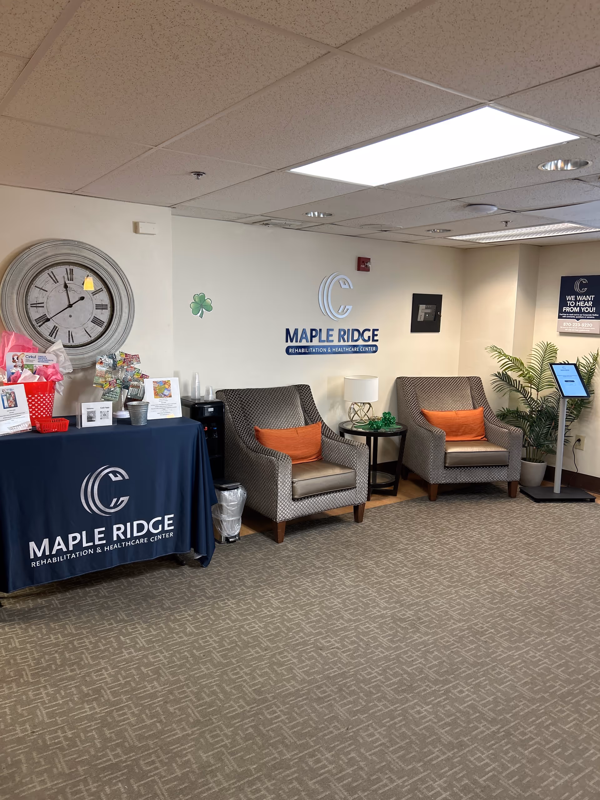 A waiting area inside Maple Ridge Rehabilitation and Healthcare Center featuring two patterned armchairs with orange cushions, a small round table with a lamp and green decoration, a large wall clock, a table covered with a Maple Ridge branded cloth holding informational materials, and a digital kiosk near a potted plant.