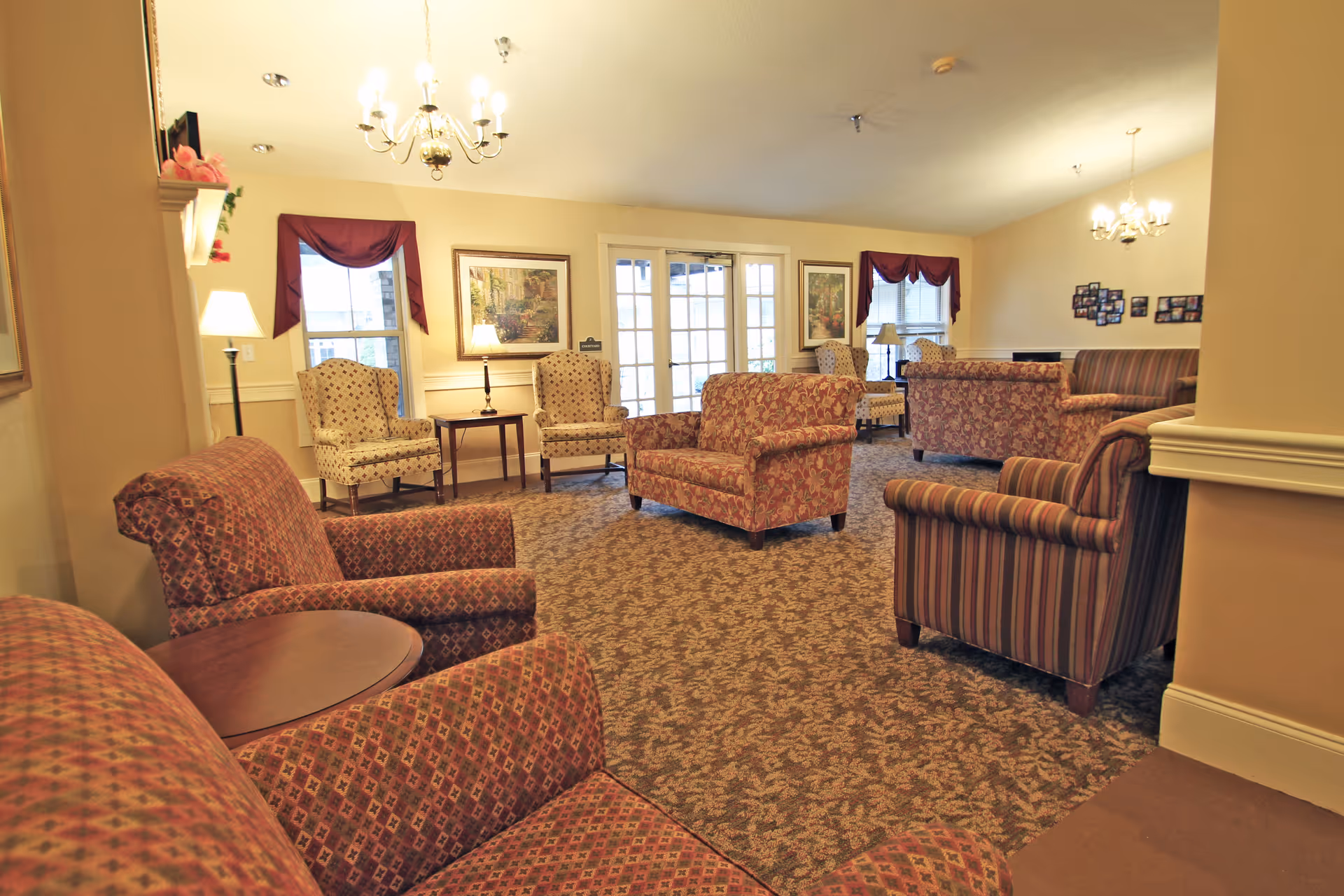 A spacious living room area in a senior living facility with multiple upholstered armchairs and sofas in patterned fabrics. The room has beige walls, carpeted floors with a floral pattern, two chandeliers, framed artwork on the walls, and windows with burgundy valances. There are also several lamps providing warm lighting and a set of double glass doors leading outside.
