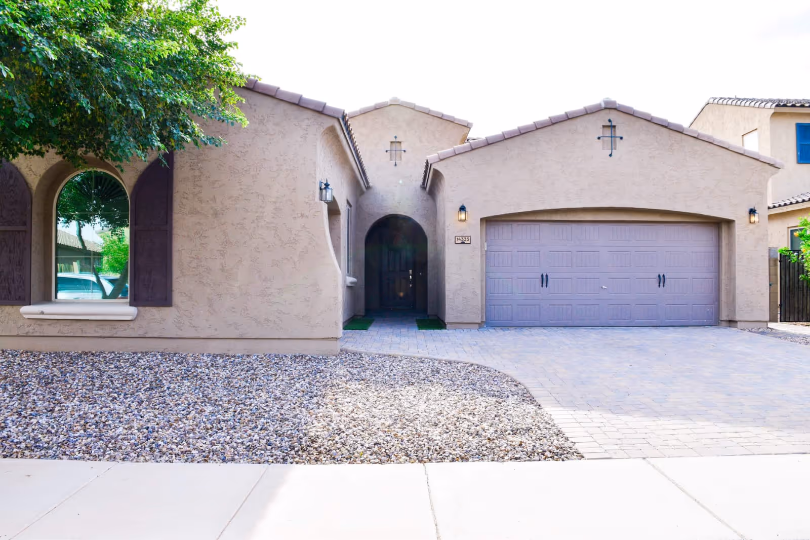 Exterior front view of a beige stucco building with a tiled roof, featuring a large garage door, an arched entryway, and a window with wooden shutters. There is a tree on the left side and a gravel landscaping area in front.