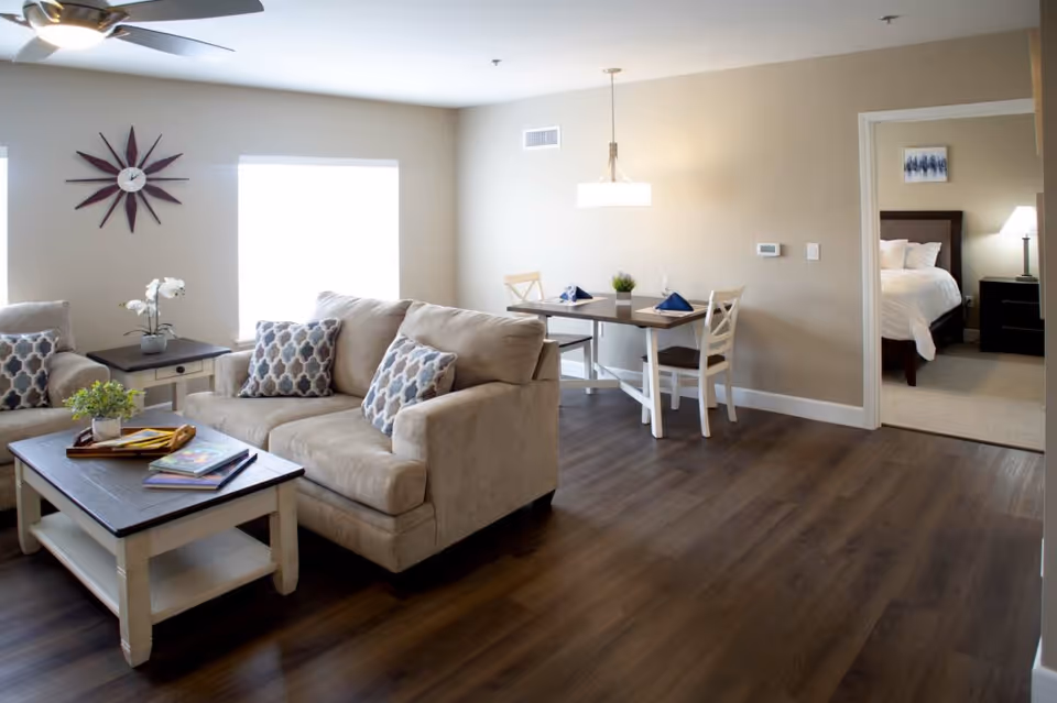 A cozy living room and dining area with beige walls and dark wood flooring. The living room features a beige sofa and armchair with patterned cushions, a wooden coffee table with books and a plant, and a side table with a white orchid. A large window with bright natural light is on the wall behind the sofa. The dining area has a small table set for two with napkins and a small plant centerpiece. A ceiling light fixture hangs above the dining table. In the background, an open doorway leads to a bedroom with a bed, nightstand, lamp, and wall art.