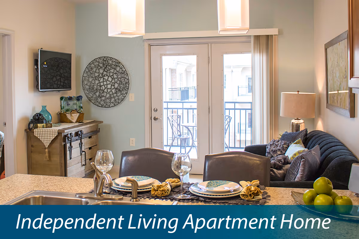 Open-plan apartment interior showing a kitchen countertop set for dining in the foreground, a sink and faucet, a sofa and TV, and glass doors opening to a balcony.