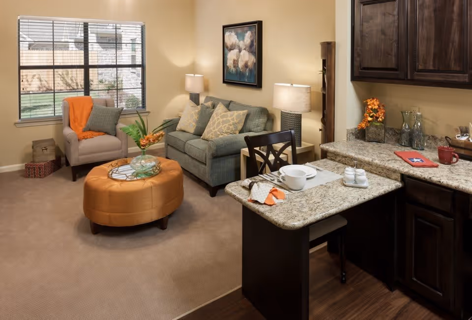 Cozy living room area with a gray sofa adorned with patterned pillows, a beige armchair with an orange throw blanket, and a round orange ottoman serving as a coffee table with a glass vase and flowers. The room has beige walls, a large window with blinds, two table lamps, and a granite countertop with a dining setup including a plate, cup, and utensils.