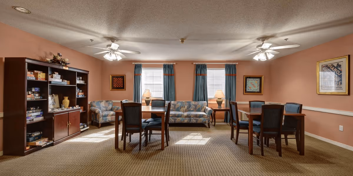 A cozy senior living common room with two wooden tables each surrounded by four chairs, a patterned sofa and armchair, two table lamps on side tables, a bookshelf filled with board games and books, and framed artwork on peach-colored walls with blue curtains on the windows.