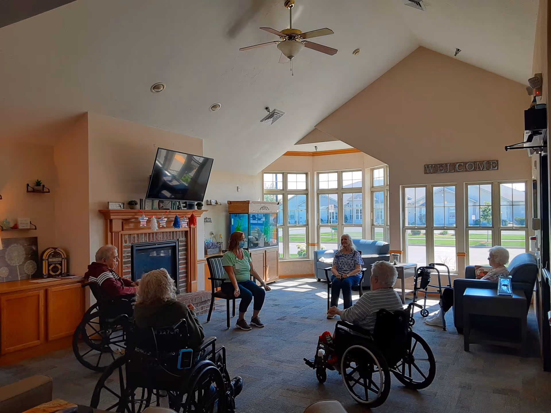 A bright common area in a senior living facility with large windows letting in natural light. Several elderly residents, some in wheelchairs, and a caregiver wearing a mask are seated in a circle. The room features a fireplace with a TV mounted above it, a ceiling fan, and comfortable seating. A 'WELCOME' sign is visible on the wall above the windows.