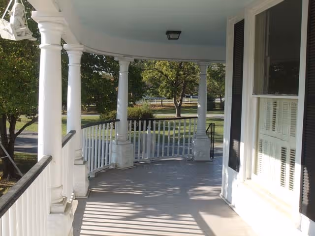 Curved covered front porch with white columns and railing overlooking a tree-lined yard.