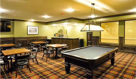 Interior view of a recreational room with a pool table in the center, several tables and chairs arranged around the room, a television mounted on the wall, and a counter area in the back. The room has patterned carpet flooring and warm lighting.