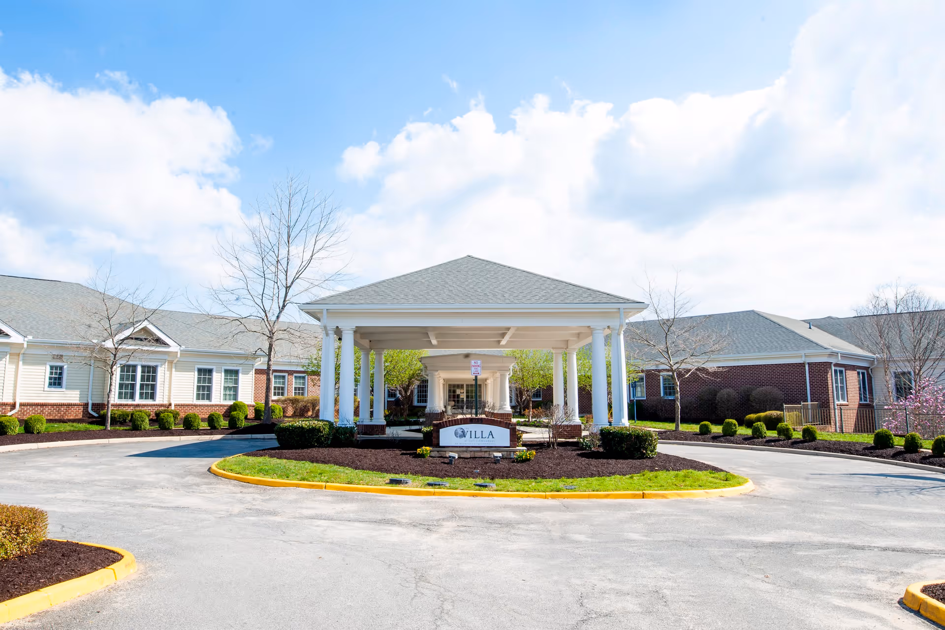 Front exterior view of The Villa at Suffield Meadows building with a covered entrance supported by white columns, surrounded by landscaped bushes and trees under a partly cloudy sky.