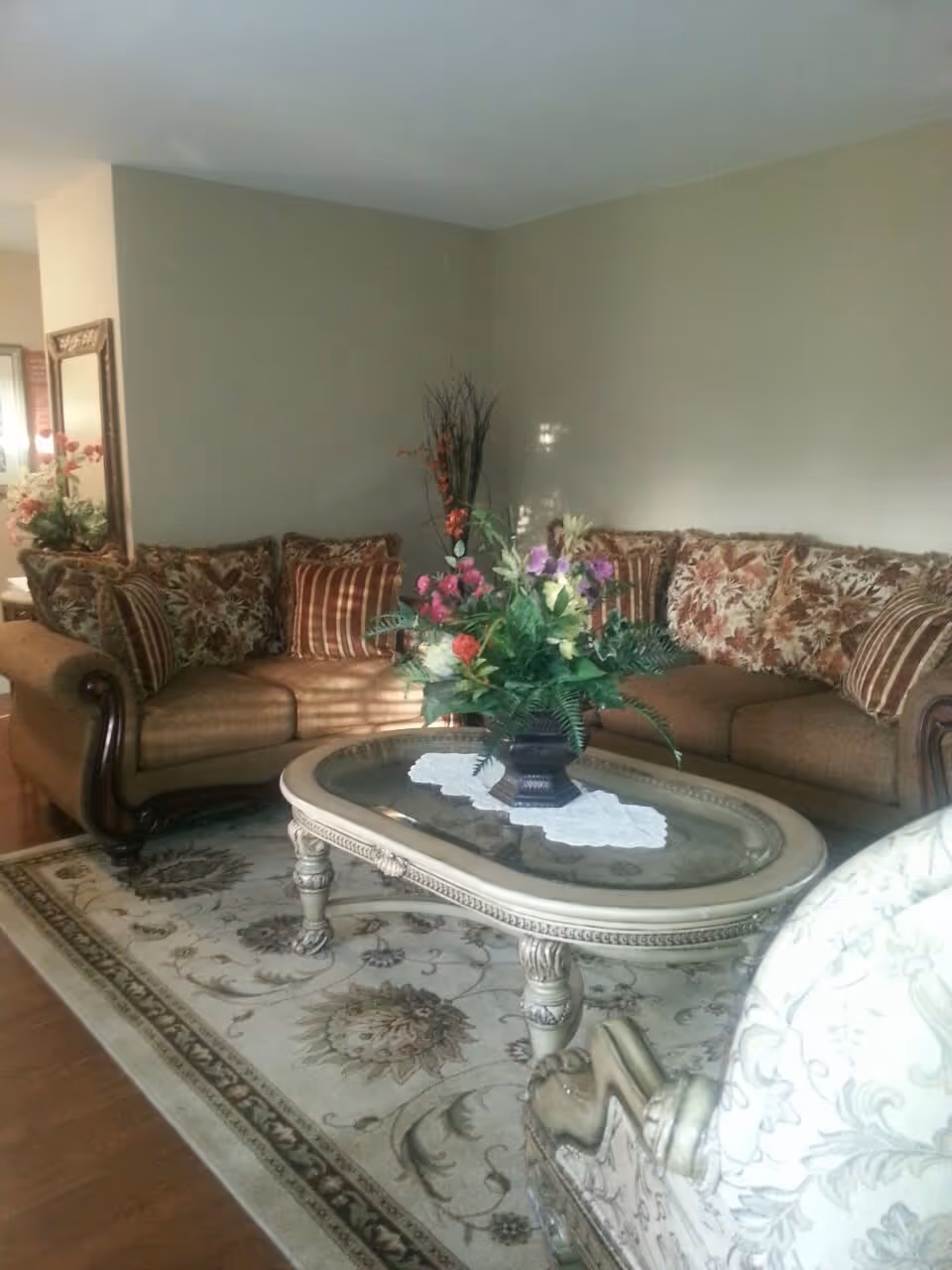 A cozy living room with two brown upholstered sofas adorned with floral and striped cushions. In front of the sofas is an ornate glass-top coffee table with a floral arrangement centerpiece. The room features a patterned area rug and a decorative mirror on the wall to the left.