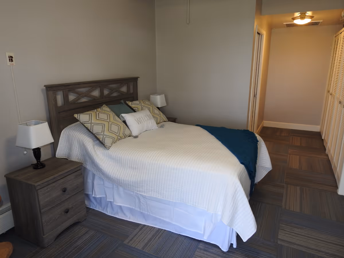 A neatly made bed with a wooden headboard in a senior living facility bedroom. The bed has a white quilt, decorative pillows including one with the word 'WELCOME', and a blue throw blanket. There are two wooden nightstands with lamps on either side of the bed. The room has gray walls and carpeted flooring with a hallway visible in the background.