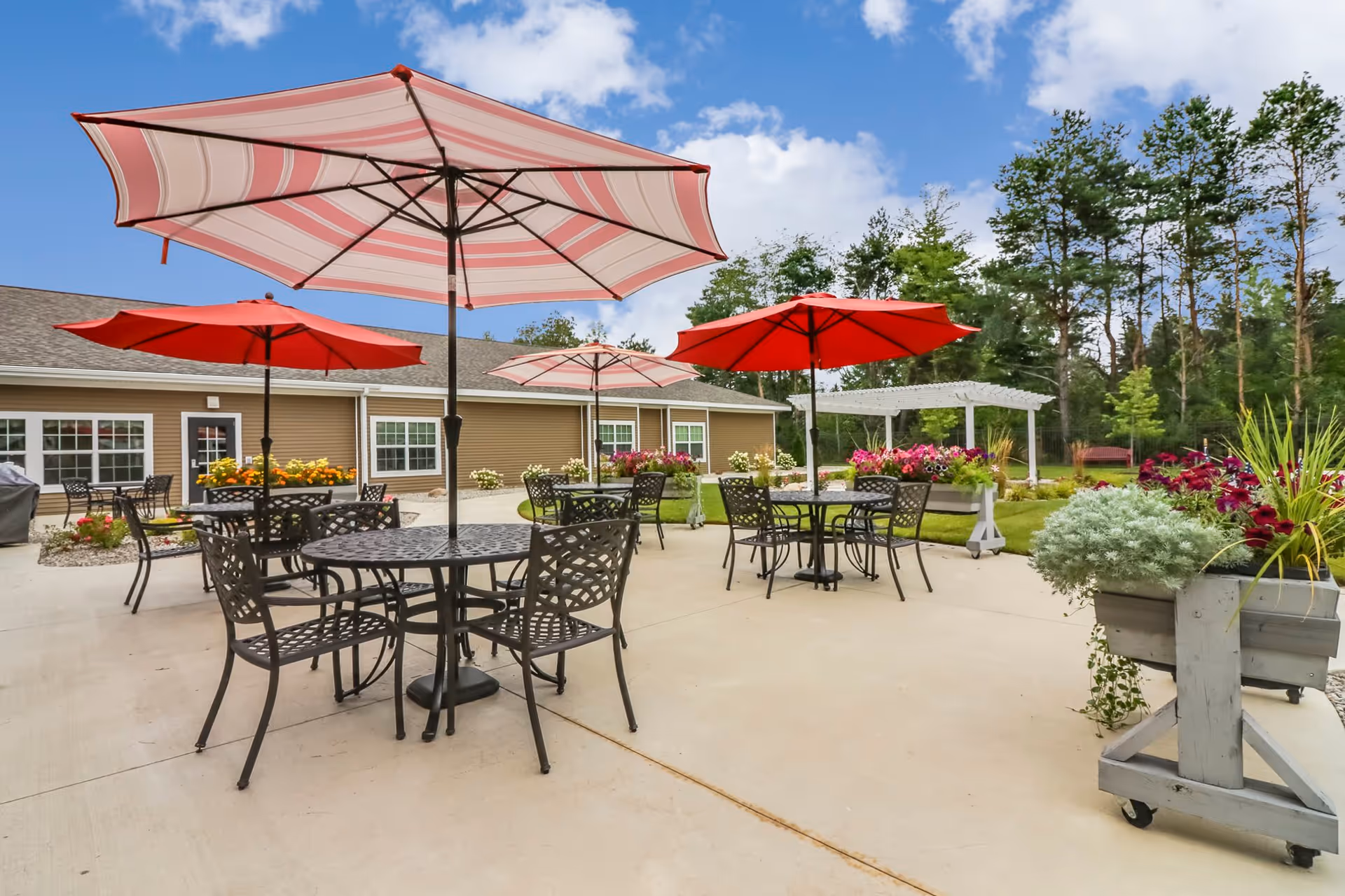 Outdoor patio area with several black metal tables and chairs, each table shaded by large red and pink striped umbrellas. The patio is surrounded by flower beds with colorful flowers and greenery, and a beige building with multiple windows is visible in the background under a partly cloudy blue sky.