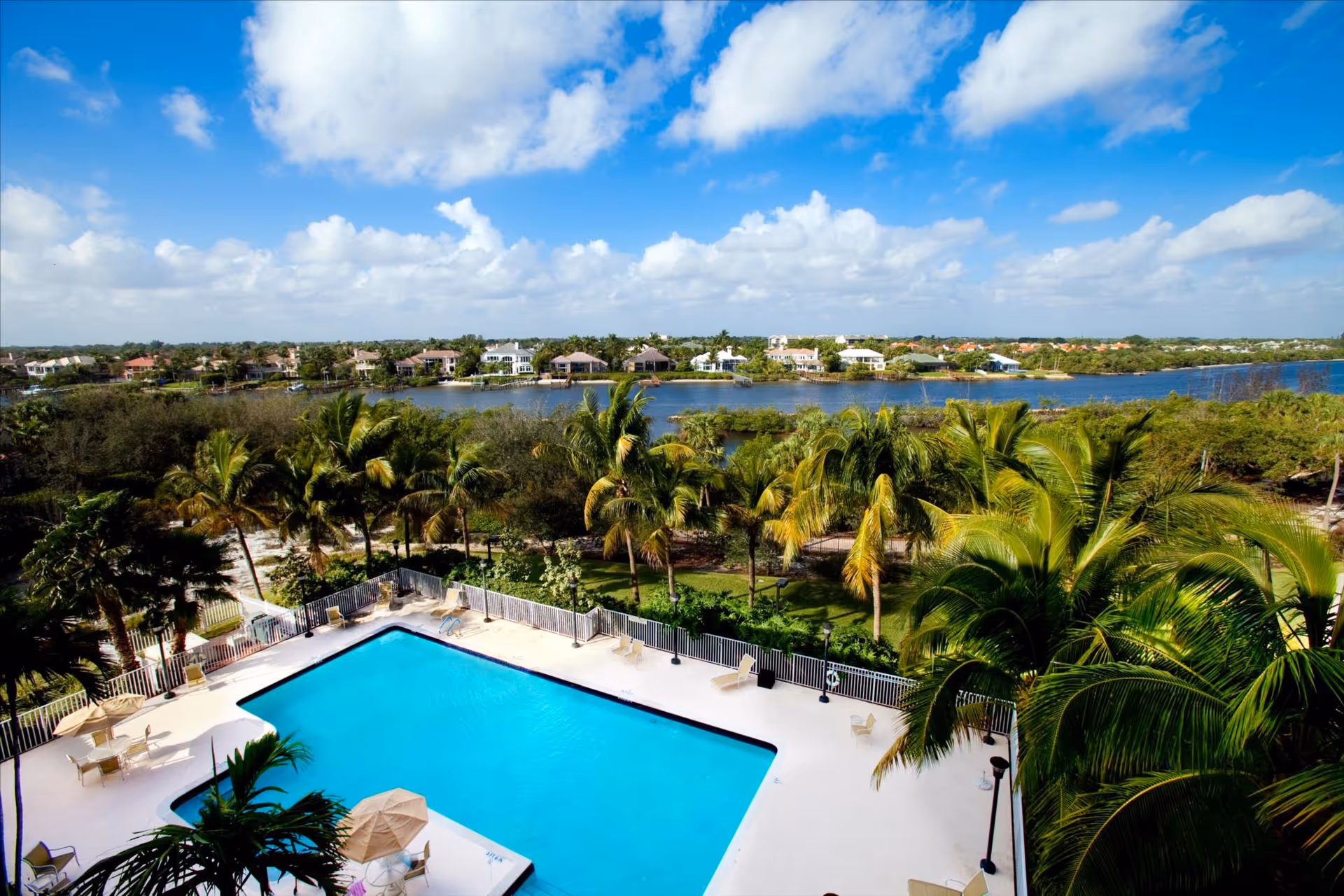 Aerial view of a swimming pool terrace with lounge chairs and umbrellas, palm trees, and a waterway with houses beyond under a blue sky.