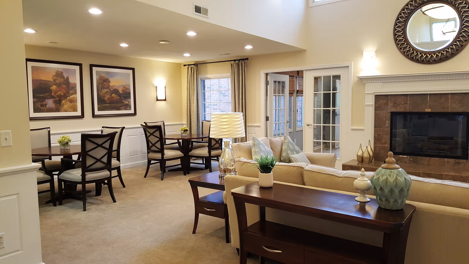 A cozy and well-lit common area in Brookfield Highlands Apartments 55+ featuring a beige sofa with decorative pillows, a wooden console table with decorative items, a fireplace with a round mirror above it, and a dining area with tables and chairs. The room has light-colored walls, carpeted floors, framed landscape paintings, and French doors leading to another room.