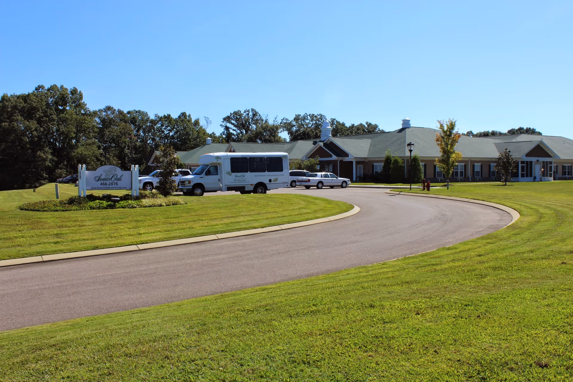 Exterior view of SomerOak Senior Living facility with a curved driveway, green lawn, several parked vehicles including a white shuttle bus, and a clear blue sky.