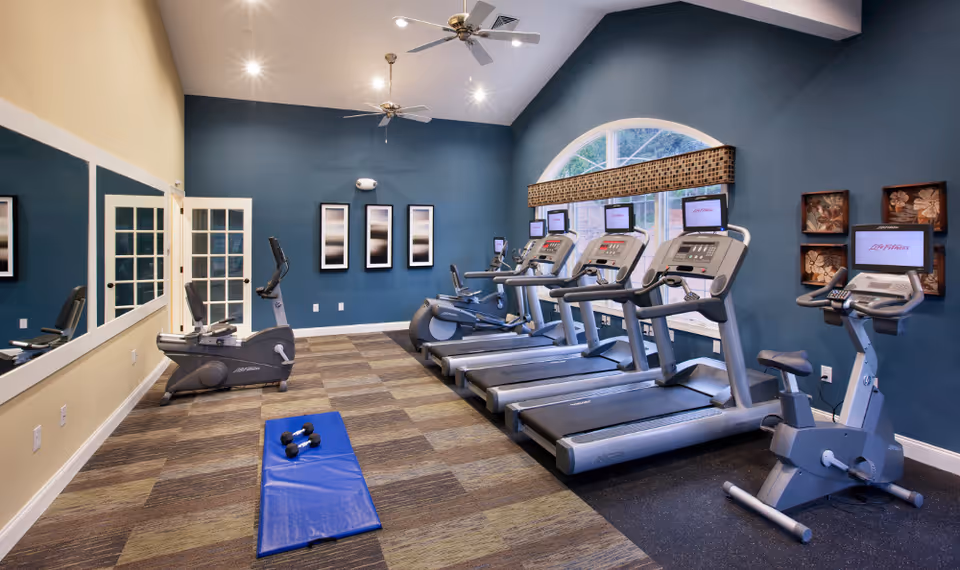 A fitness room with several treadmills, stationary bikes, a blue exercise mat with dumbbells on it, large wall mirror, and blue walls with framed artwork. The room has a large arched window with a patterned valance and ceiling fans.