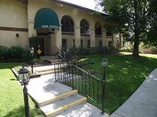 Exterior view of a two-story building with arched windows and a green awning labeled 'Casa Glendale' over the entrance. There is a pathway with steps and black metal railings leading up to the entrance, surrounded by green grass and trees.