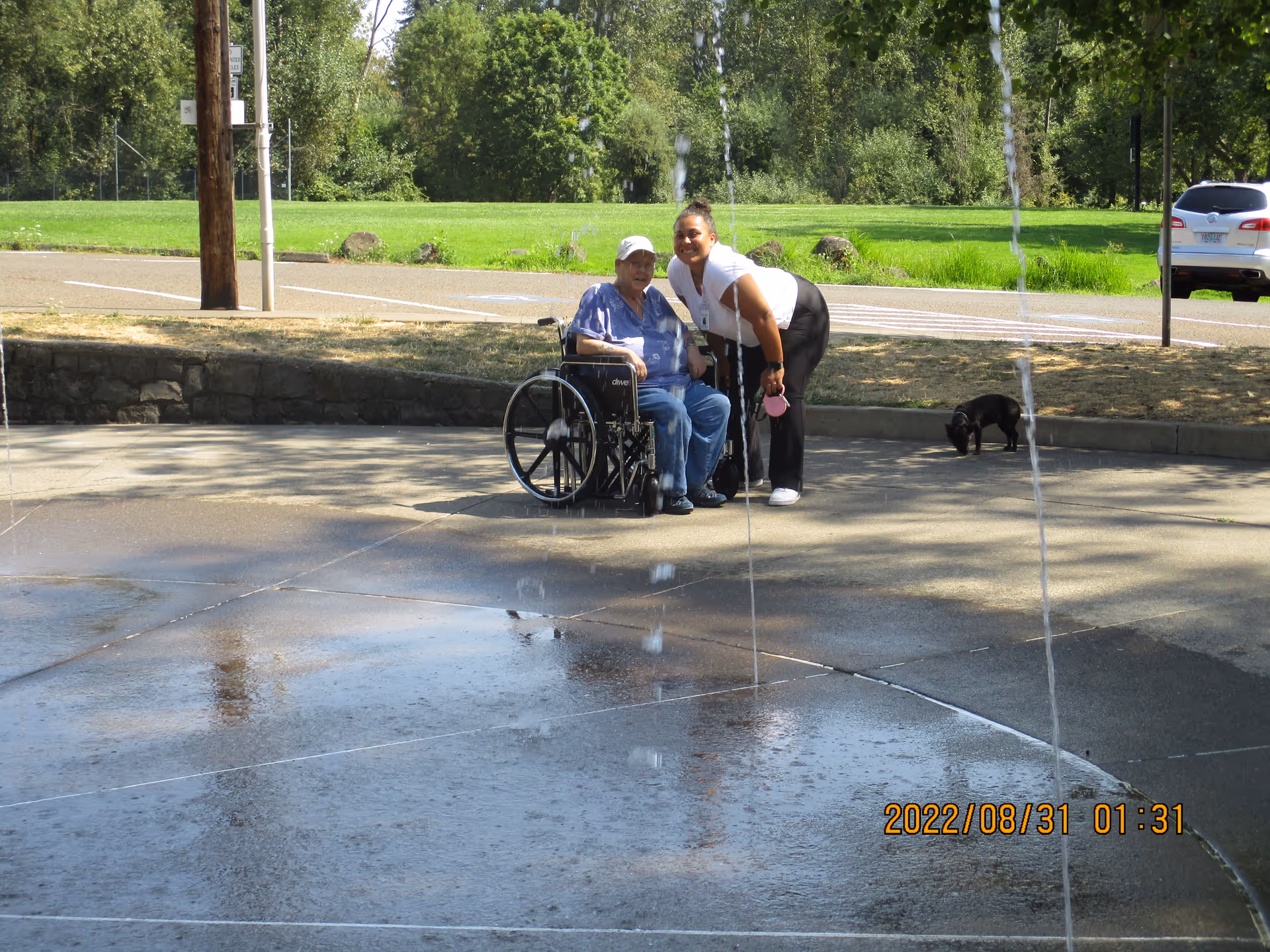 An elderly man in a wheelchair and a woman standing beside him smiling outdoors near a water fountain feature with streams of water shooting up from the ground. There is a small black dog sniffing the ground nearby, and green grass and trees are visible in the background.