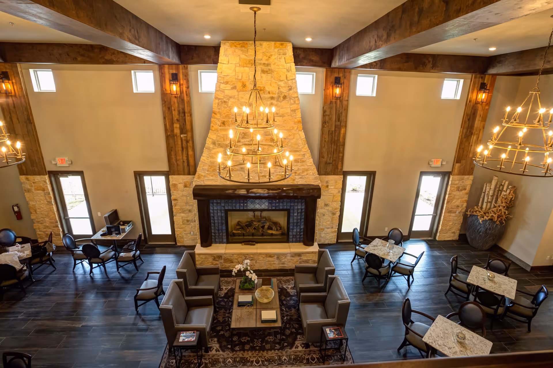 Spacious common area with a large stone fireplace in the center, surrounded by comfortable seating including armchairs and small tables with chairs. The room features high ceilings with exposed wooden beams, multiple windows, and two large chandeliers providing warm lighting.