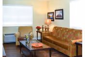 A cozy living room with a patterned orange sofa, a striped armchair, a wooden coffee table with decorative items, a side table with a lamp, and two framed pictures on the wall. A window with closed blinds and an air conditioning unit is visible.