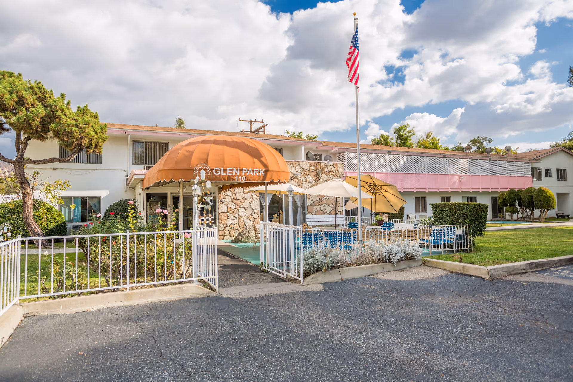 Front entrance of the Glen Park senior living building with an orange awning, stone facade, American flag, and outdoor seating area.