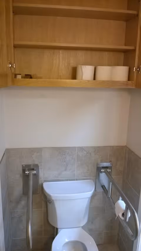 A bathroom with a white toilet positioned against a tiled wall. There are two metal grab bars on either side of the toilet, one of which has a toilet paper roll holder with a roll of toilet paper. Above the toilet is a wooden cabinet with open doors, showing some toilet paper rolls and other small items inside.