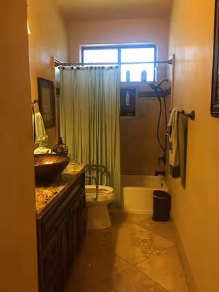 A bathroom with beige walls and tiled floor featuring a wooden vanity with a vessel sink and a decorative faucet on the left, a toilet in the center, and a bathtub with a shower curtain and showerhead on the right. A window above the bathtub lets in natural light. Towels hang on the wall to the right and a small trash bin is next to the bathtub.