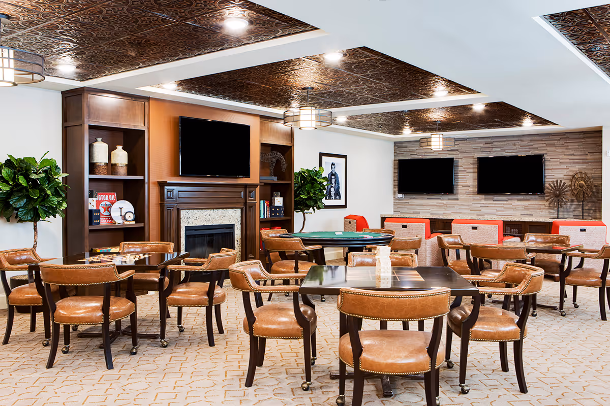A spacious common area with multiple tables and leather chairs arranged for group activities. The room features a decorative ceiling, a fireplace with a mounted TV above it, two additional TVs on a stone accent wall, and various decorative items on shelves. There are also two large potted plants and framed artwork on the walls.