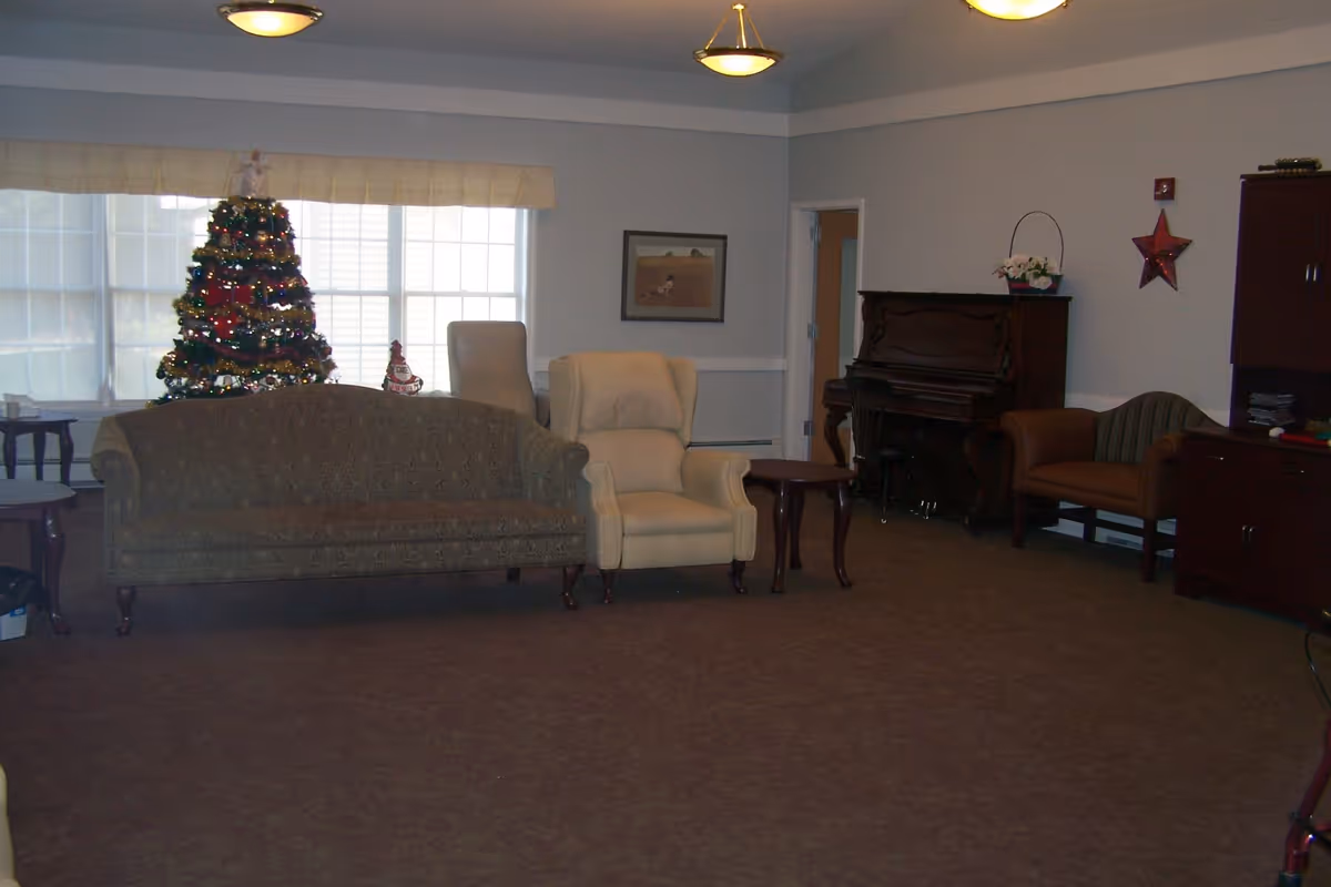 A cozy living room with a decorated Christmas tree near a large window, a patterned sofa, a beige armchair, a small wooden side table, a piano against the wall, and additional seating including a striped armchair. The room has soft lighting from ceiling fixtures and neutral-colored walls.