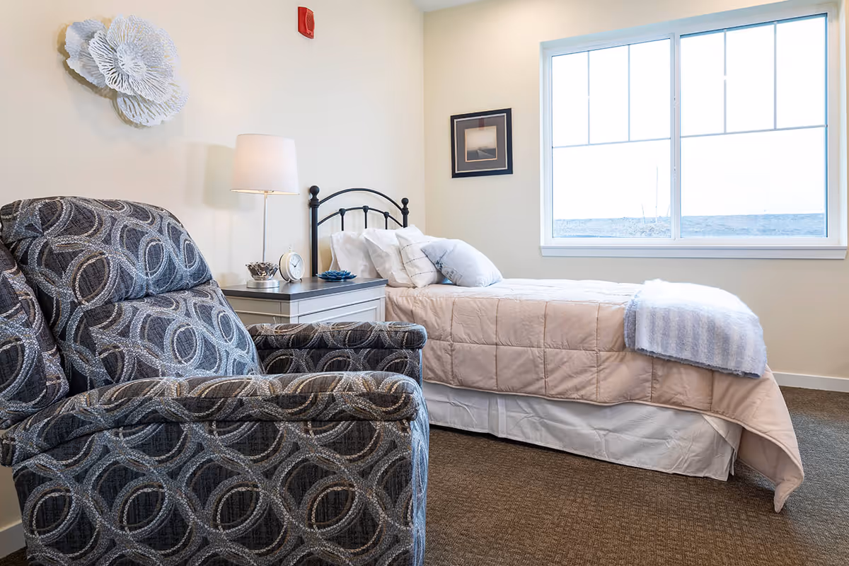 A cozy assisted-living bedroom with a single bed, nightstand and lamp, and a patterned recliner beside a large window.