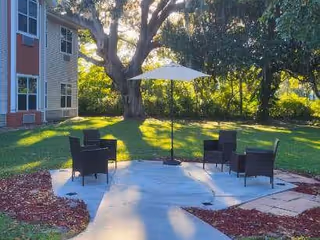 Outdoor seating area with four black wicker chairs arranged around a white umbrella on a concrete patio, surrounded by green grass, trees, and a building on the left side.