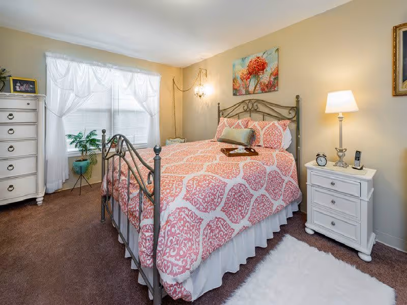 A cozy bedroom with a metal frame bed dressed in a pink and white patterned comforter and pillows. There is a white nightstand with a lamp, alarm clock, and phone on the right side of the bed. A white dresser with drawers is on the left side near a window with sheer white curtains. A green potted plant is placed near the window. The walls are painted beige with a floral painting hanging above the bed. The floor is carpeted in brown with a white rug beside the bed.