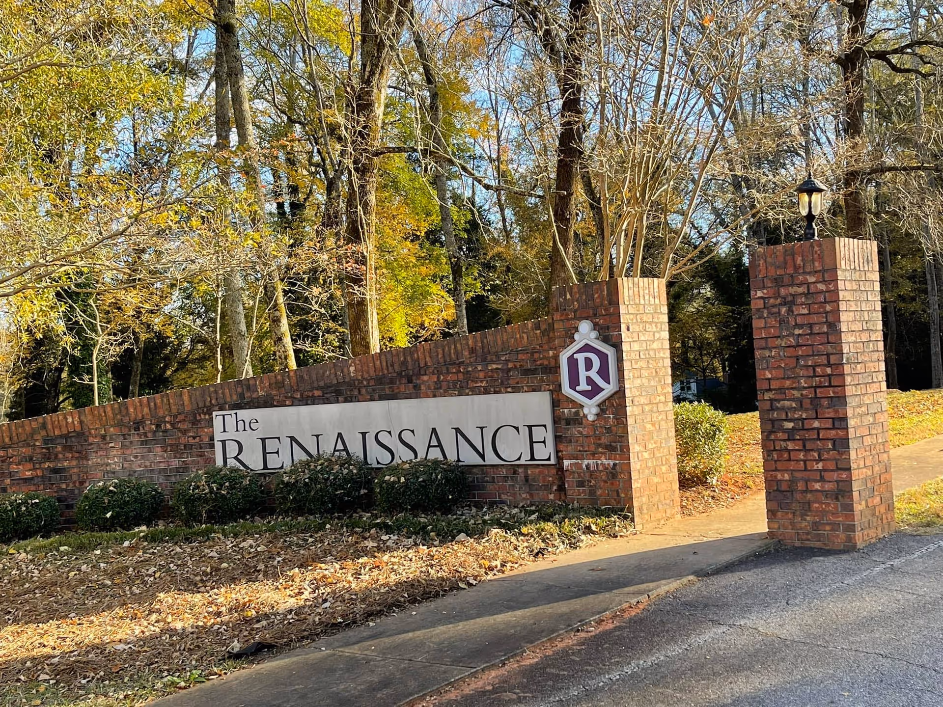 Brick entrance sign for The Renaissance Senior Living facility surrounded by trees with autumn foliage and a paved driveway.