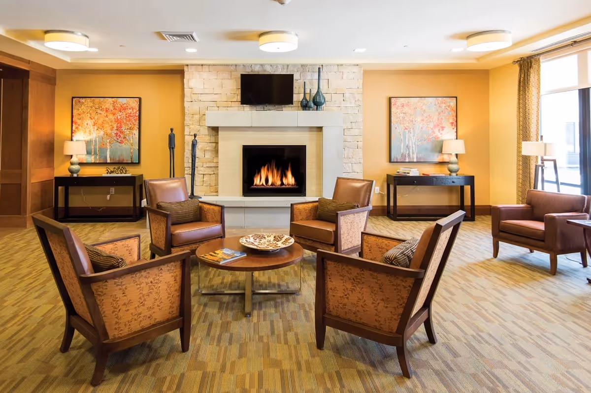 A cozy living room area with a stone fireplace and a flat-screen TV mounted above it. Four wooden armchairs with brown cushions are arranged around a round wooden coffee table with a decorative bowl and a book. The room has warm yellow walls, two side tables with lamps and artwork featuring autumn trees, and large windows with curtains letting in natural light.