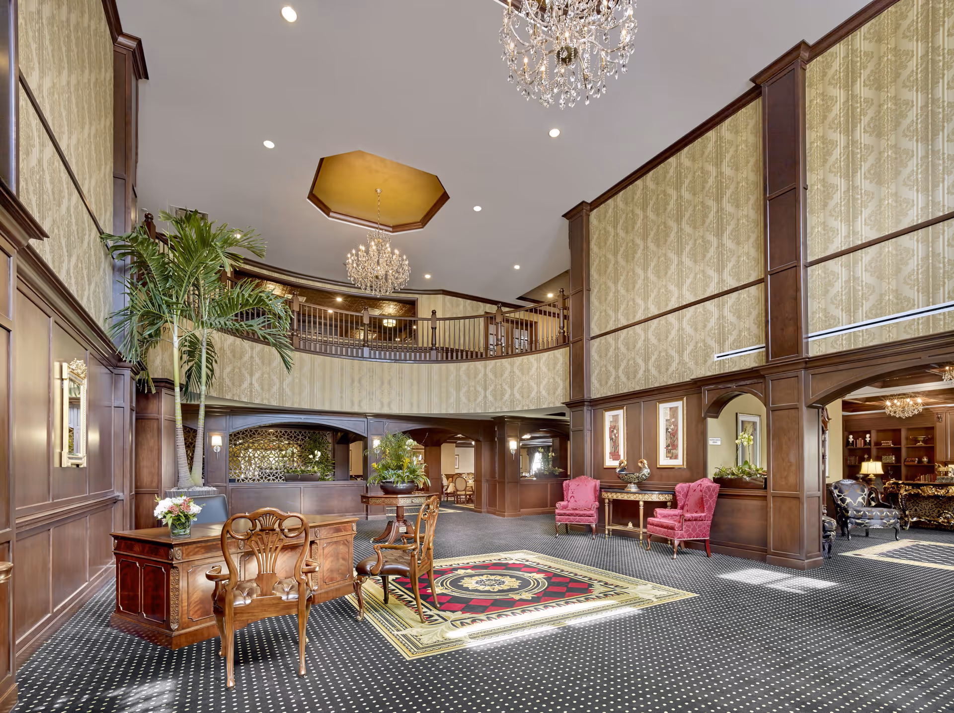 Spacious and elegant lobby area with high ceilings, chandeliers, wood paneling, patterned carpet, and classic furniture including a wooden desk with chairs, two pink armchairs, and decorative plants.