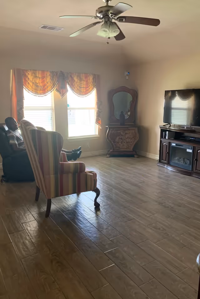A living room with wooden flooring, a ceiling fan, two windows with orange and yellow curtains, a striped armchair, a person sitting in a recliner chair, a decorative wooden dresser with a mirror, and a flat-screen TV on a wooden TV stand.