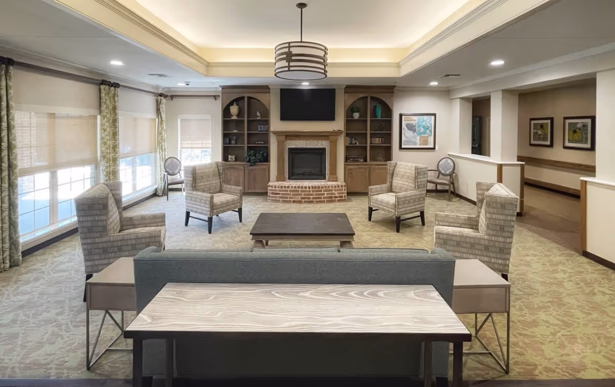 A spacious living room with a central fireplace and a mounted TV above it. The room features four patterned armchairs arranged around a low coffee table, a sofa with a wooden table behind it, large windows with green patterned curtains, built-in wooden shelves flanking the fireplace, and framed artwork on the walls. The ceiling has recessed lighting and a modern circular chandelier.