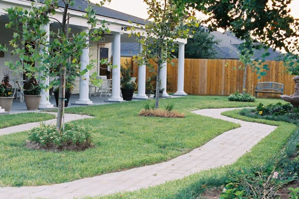 A landscaped outdoor area with a winding paved walkway, green grass, small trees, and flower beds. There is a building with white columns and a porch with chairs in the background, along with a wooden fence and a bench near the back right side.